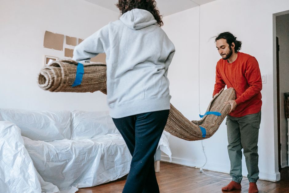A couple unrolls a carpet in their new apartment, amidst moving and unpacking.